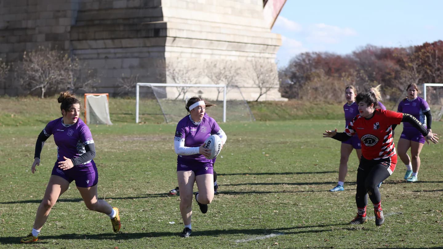 Women's rugby players carrying the ball beneath the Robert F. Kennedy Bridge